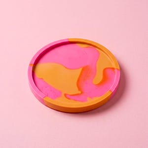 Peut inclure: Un dessous de verre circulaire présentant un motif marbré aux couleurs rose vif et orange éclatant. Le dessous de verre possède un rebord extérieur surélevé et une surface lisse et plate, affichant un design tourbillonnant et mélangé.