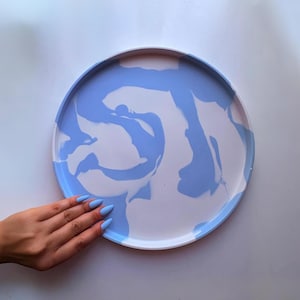 Peut inclure: Un plateau décoratif rond avec un motif marbré dans les tons bleu clair et blanc. Le plateau a un bord surélevé et une surface lisse. Le plateau est présenté sur un fond blanc.