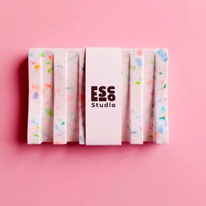 Puede incluir: Jabonera rectangular con diseño moteado en tonos rosa, azul, verde y amarillo. Tiene ranuras verticales y una banda blanca con el logotipo "ESS Studio". El fondo es de color rosa liso.