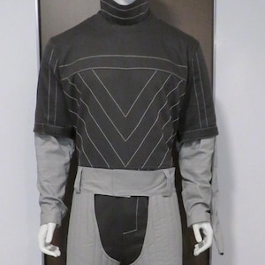 Può includere: Un manichino indossa un completo in stile futuristico. L'ensemble include una maglia a collo alto grigio scuro con cuciture bianche, maniche grigio chiaro, una cintura abbinata e pantaloncini trapuntati. L'outfit è completato da pantaloni grigio scuro.