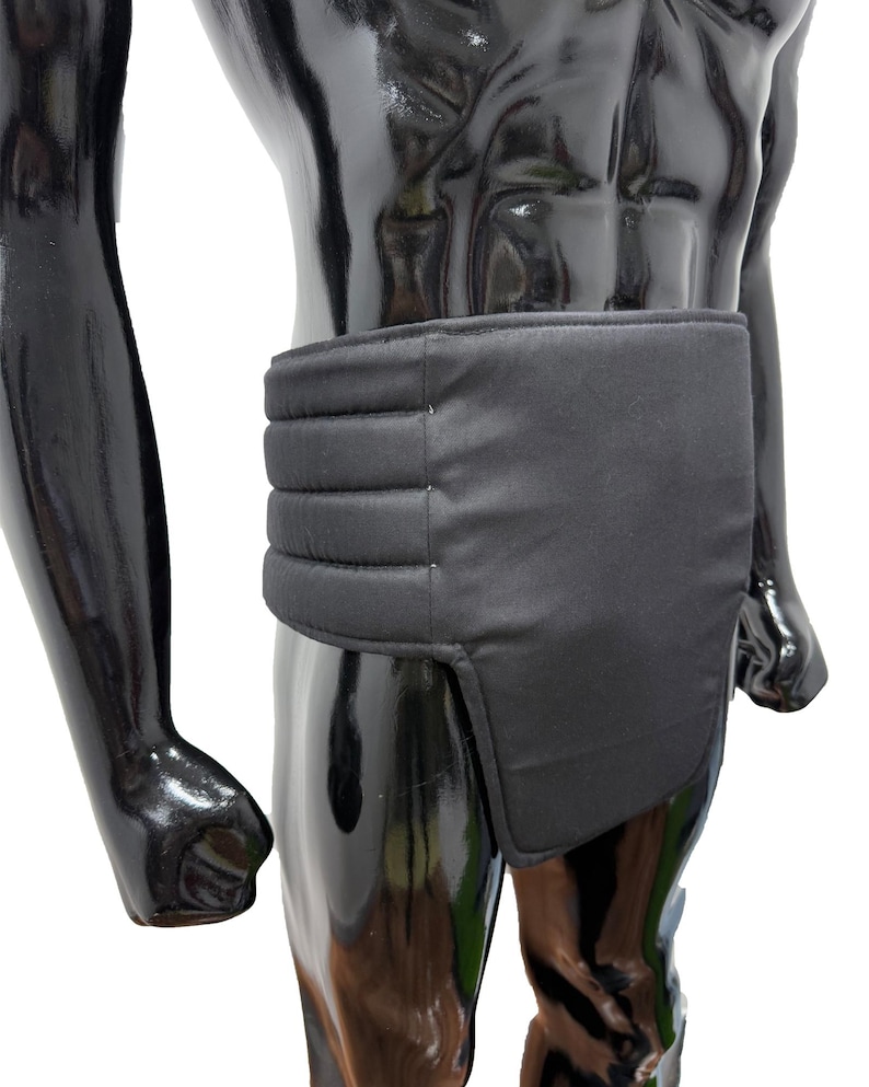 May include: A black garment with a padded, horizontal ribbed design across the midsection. The garment appears to be worn around the waist and hips, with a rectangular flap extending down the front. The mannequin is black and glossy.