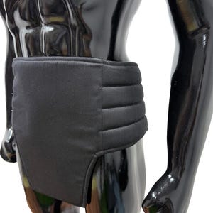 May include: A black support garment, likely for the lower back or abdomen, is displayed on a mannequin. The garment has a rectangular shape with a padded, ribbed section across the midsection. The material appears to be a matte, non-reflective fabric.