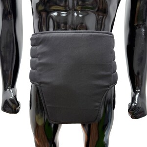 May include: Black padded support garment. The garment is worn around the waist and lower abdomen. The support has a rectangular shape with horizontal stitching across the back. The garment is displayed on a black mannequin.