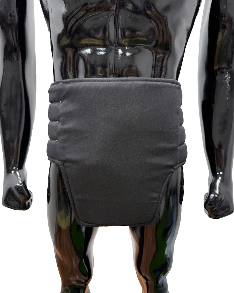 May include: Black padded support garment. The garment is worn around the waist and lower abdomen. The support has a rectangular shape with horizontal stitching across the back. The garment is displayed on a black mannequin.