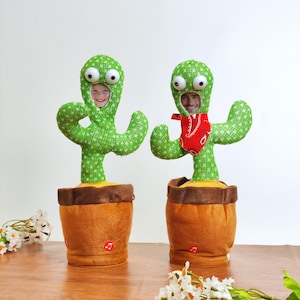 May include: Two green cactus-shaped plush toys with faces in the center, set in brown pots. One cactus has a red bandana. The toys have large, googly eyes and are on a wooden surface. The cactus toys are a fun, novelty item.