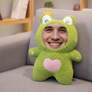 May include: A green plush frog-shaped pillow with a face cutout. The pillow has a pink heart on its chest and a small yellow crown. The person's face is visible through the cutout. The pillow is on a gray couch.