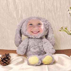 Könnte beinhalten: Ein flauschiges, graues Hasenkostüm mit dem Gesicht eines Kindes in der Mitte. Der Hase hat lange, schlaffe Ohren, einen flauschigen Körper und gelbe Fußpolster. Ein Tannenzapfen liegt links vom Hasen auf einer satinartigen Oberfläche.