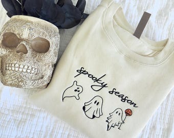 May include: A cream-colored crewneck sweatshirt featuring black embroidered text that reads spooky season, accompanied by three small ghost figures, one of which is holding a small orange jack-o'-lantern balloon on a thin black string.