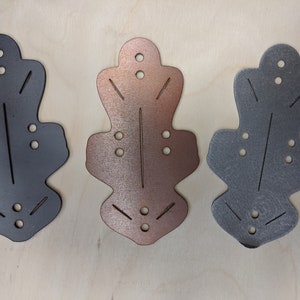 Op de afbeelding: Drie metalen decoratieve bladvormige uitsparingen in verschillende kleuren: donkergrijs, koper en lichtgrijs. Elke uitsparing heeft kleine gaatjes en geëtste lijnen, met een afgeronde bovenkant en een uitlopende basis. Ze liggen op een licht houten oppervlak.