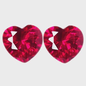 Könnte beinhalten: Zwei herzförmige, facettierte rote Edelsteine im Brillantschliff. Jeder Stein weist einen tiefen karmesinroten Farbton und mehrere dreieckige Facetten auf, die das Licht reflektieren und ein funkelndes Aussehen mit scharfen, definierten Kanten erzeugen.