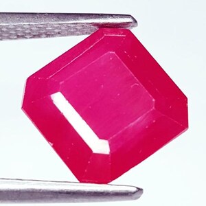 May include: A vibrant, deep pinkish-red gemstone with a faceted, rectangular emerald cut. The stone is held by a pair of metallic tweezers, showcasing its translucent quality and sharp, clean edges against a plain, bright white background.