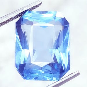 May include: A light blue, rectangular-cut gemstone, held by silver tweezers. The gem's facets are precisely cut, reflecting light and creating a sparkling effect. The stone is a light shade of blue. The tweezers are holding the gem.