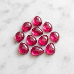 May include: A collection of ten loose, teardrop-shaped gemstones in a vibrant, deep magenta-pink hue. Each stone features a smooth, polished cabochon surface with a rounded top and a pointed base, displaying a translucent quality and consistent colour.