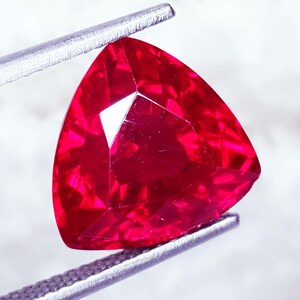 Könnte beinhalten: Ein leuchtend tiefroter Edelstein in dreieckiger Form mit zahlreichen Facetten, die das Licht reflektieren. Der Stein wird von einer silberfarbenen Metallpinzette gehalten, was seine Transparenz und die intensive karmesinrote Farbe hervorhebt.