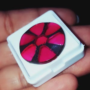 Puede incluir: Un objeto decorativo redondo con un vibrante diseño rojo en forma de pétalos sobre un fondo negro brillante. Se presenta en un contenedor cuadrado blanco, lo que sugiere que es una piedra preciosa o un elemento decorativo.