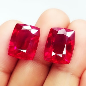 Puede incluir: Dos gemas facetadas, de talla cojín, de color rojo intenso. Las gemas tienen un alto brillo y se sostienen en una mano sobre un fondo blanco. Las gemas son de un color rojo vibrante y saturado.