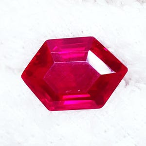Op de afbeelding: Een gefacetteerde, zeshoekige edelsteen in een levendige rode tint. De edelsteen heeft meerdere facetten die licht reflecteren, waardoor een sprankelend effect ontstaat. De edelsteen ligt op een witte, getextureerde achtergrond.