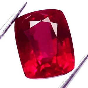 May include: A vivid, cushion-cut ruby gemstone, held by silver tweezers. The gem is a deep, saturated red with internal facets that reflect light. The shape is a square with rounded corners, showcasing the stone's brilliance.