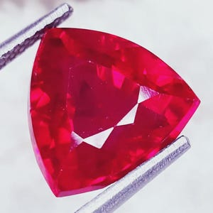 Peut inclure: Un rubis taillé en triangle, maintenu par une pince argentée. La gemme est d'un rouge profond avec des facettes qui captent la lumière, créant des reflets internes brillants. La forme est un triangle équilatéral parfait.