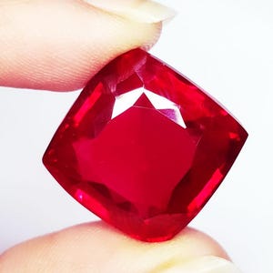 Puede incluir: Una gema de color rojo intenso y vibrante con talla cojín, que presenta múltiples facetas triangulares y trapezoidales que reflejan la luz. La piedra tiene una forma cuadrada con esquinas redondeadas y un color carmesí saturado y translúcido.