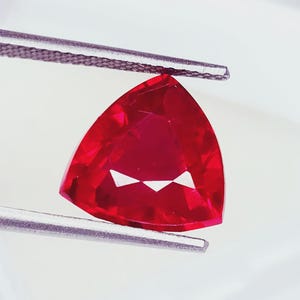 May include: A vibrant, deep red gemstone cut into a triangular shape with multiple facets that reflect light. The gemstone is held securely between the two metal tips of a pair of silver-colored tweezers, showcasing its clarity and color.