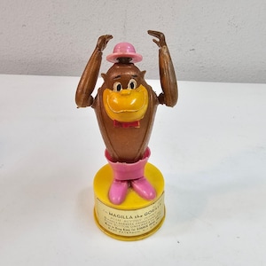 May include: A vintage Magilla Gorilla push button toy. The brown gorilla figure has a yellow beak, pink bow tie, and a pink hat. The base is yellow with pink boots and the text "I'm Magilla the Gorilla".