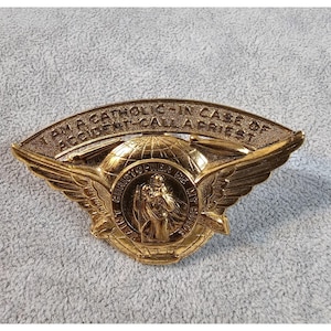 Könnte beinhalten: Eine goldfarbene religiöse Anstecknadel des Heiligen Christophorus. Die Anstecknadel zeigt einen Globus, Flügel und das Bild des Heiligen Christophorus, der ein Kind trägt. Der Text lautet "SAINT CHRISTOPHER BE MY GUIDE" und "I AM A CATHOLIC - IN CASE OF ACCIDENT - CALL A PRIEST."