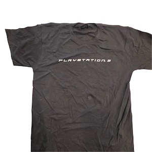May include: A dark gray short-sleeve t-shirt featuring the white text PLAYSTATION 3 printed across the center of the chest. The garment has a standard crew neckline and a relaxed fit, showcasing a simple design with a prominent brand logo.