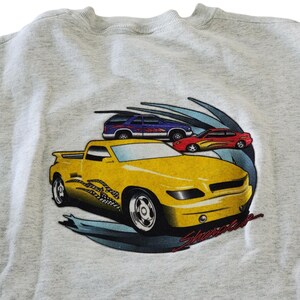Könnte beinhalten: Ein hellgraues Sweatshirt mit einer Grafik von drei Autos: einem gelben Pickup-Truck, einem roten Sportwagen und einem blauen SUV. Der gelbe Truck hat ein kariertes Flaggen-Design. Das Wort "Showtime" ist in roter Schrift geschrieben.