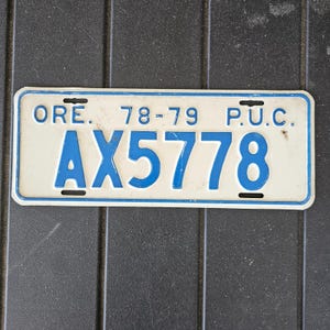 May include: Vintage Oregon license plate from 1978-79, featuring the plate number AX5778 in blue against a white background. The plate has the letters ORE. and P.U.C. at the top, with a blue border and black mounting slots.