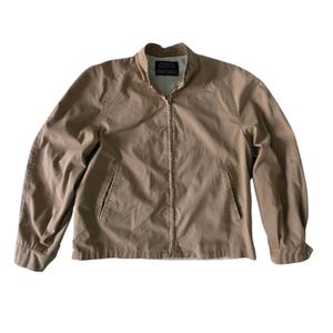 May include: A tan zip-up jacket featuring a stand-up collar, long sleeves with buttoned cuffs, and two slanted front pockets. The garment has a central zip closure and a visible interior label at the neckline, showcasing a simple, structured design.