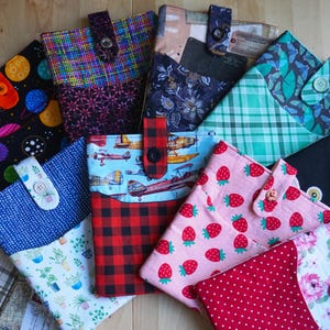 May include: A selection of fabric book covers with diverse patterns. Designs include planets, floral, plaid, strawberries, and aeroplanes. Each cover has a button closure. The covers are in shades of blue, red, pink, and green.