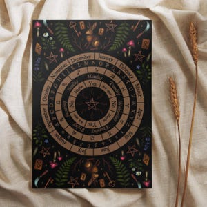 May include: A black circular divination board featuring a central pentagram, concentric rings with days of the week, months, letters, and zodiac symbols. The design includes illustrations of rabbits, mushrooms, ferns, keys, and runes in earthy tones.