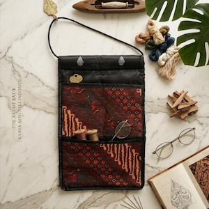 May include: A black and red batik fabric organizer bag with a shoulder strap, holding spools of thread and eyeglasses. The image also includes a wooden shuttle, needles, and a book with illustrations. The text "The Art of Batik" is visible.