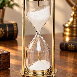 May include: An hourglass featuring a polished gold-tone metal frame with cylindrical pillars and circular top and bottom plates. The clear glass bulb contains fine white sand, with a portion of the sand currently flowing into the bottom chamber.