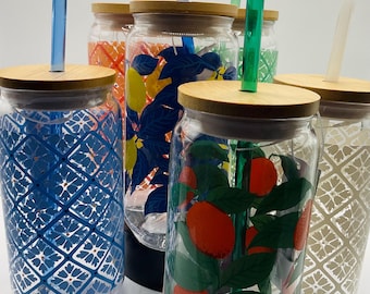 May include: A collection of clear glass drinking jars with bamboo lids and colorful straws. The jars feature various designs, including blue geometric patterns, orange citrus fruits, and floral motifs. Each jar is approximately 6 inches tall.