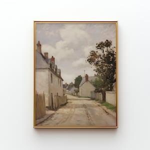 May include: A framed landscape painting depicts a quaint village street. White buildings with gray roofs line the road, with a cloudy sky overhead. The artwork is in a gold frame, adding a touch of elegance to the scene.