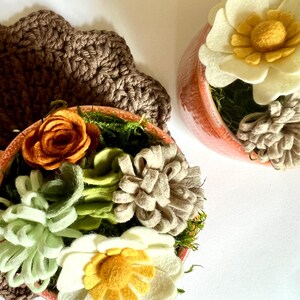 Op de afbeelding: Twee kleine terracotta potten gevuld met vilten bloemen en groen. E&eacute;n pot heeft een grote witte en gele bloem, terwijl de andere een oranje roos en diverse vilten vetplanten bevat. Op de achtergrond een bruine gehaakte onderzetter.