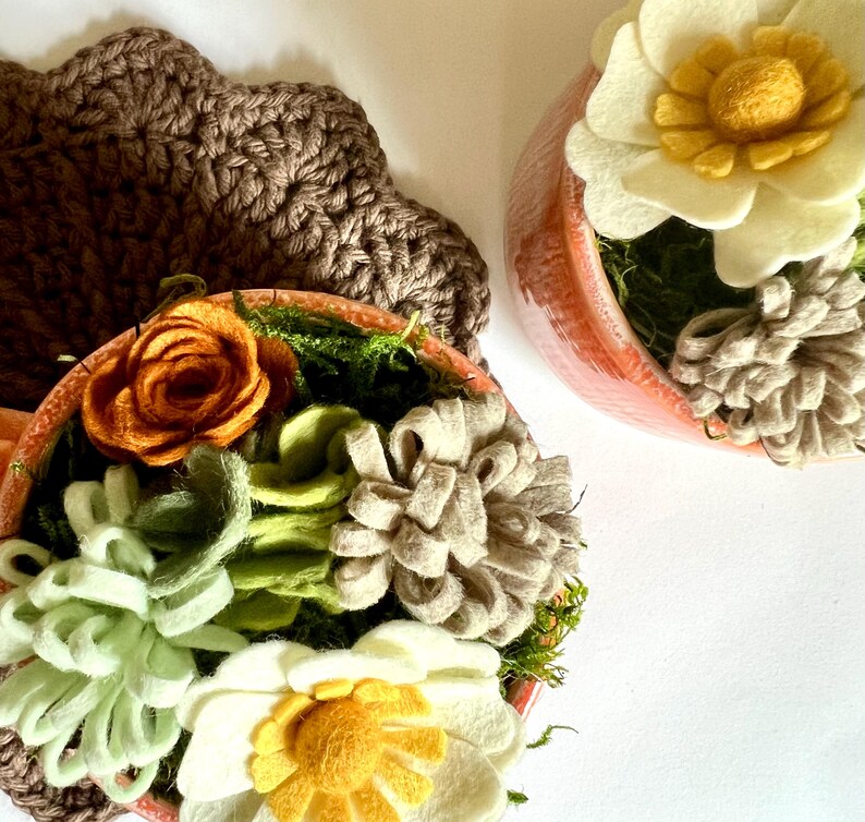 Op de afbeelding: Twee kleine terracotta potten gevuld met vilten bloemen en groen. E&eacute;n pot heeft een grote witte en gele bloem, terwijl de andere een oranje roos en diverse vilten vetplanten bevat. Op de achtergrond een bruine gehaakte onderzetter.