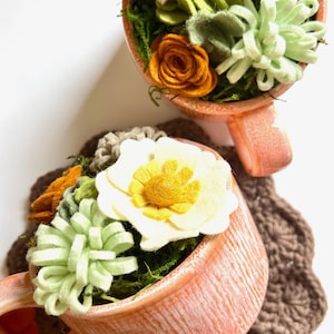 Op de afbeelding: Twee perzikroze theekopjes gevuld met vilten bloemen en groen. De bloemen zijn onder meer witte, gele en oranje rozen en vetplanten. De kopjes staan op een bruine gehaakte onderzetter en een witte achtergrond.