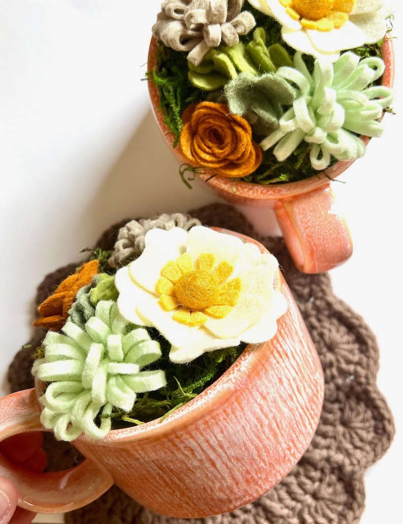 Op de afbeelding: Twee perzikroze theekopjes gevuld met vilten bloemen en groen. De bloemen zijn onder meer witte, gele en oranje rozen en vetplanten. De kopjes staan op een bruine gehaakte onderzetter en een witte achtergrond.