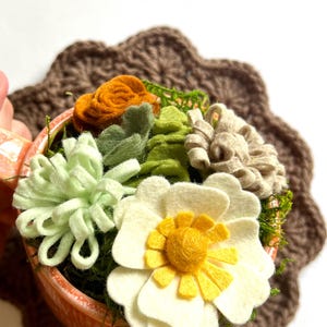 Op de afbeelding: Een viltarrangement van vetplanten in een oranje keramische beker. Het arrangement bevat een grote witte bloem met een geel hart, groene en bruine vilt vetplanten en oranje vilt rozen. De beker staat op een bruine gehaakte onderzetter.