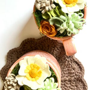 Op de afbeelding: Twee decoratieve theekopjes gevuld met vilten bloemen in tinten wit, geel, oranje en groen, geplaatst op een bruine gehaakte onderzetter. De theekopjes hebben een perzikkleurige buitenkant en zijn gevuld met mos.