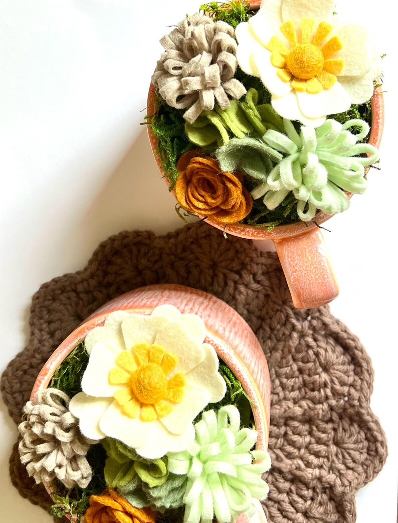 Op de afbeelding: Twee decoratieve theekopjes gevuld met vilten bloemen in tinten wit, geel, oranje en groen, geplaatst op een bruine gehaakte onderzetter. De theekopjes hebben een perzikkleurige buitenkant en zijn gevuld met mos.