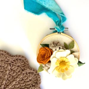 May include: An embroidered floral hoop art piece featuring a cream-colored background with felt flowers in shades of orange, white, and yellow. The hoop is suspended by a turquoise ribbon. A brown crocheted item is in the lower left corner.