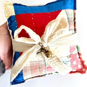May include: A small, square patchwork pillow with a cream-colored ribbon tied around it. The pillow features red, blue, and patterned fabric squares. A small charm with a key and bell hangs from the ribbon. The pillow is held by a person.