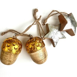 May include: Two wooden acorn ornaments with metallic gold caps and two wooden star ornaments with metallic silver finishes. Each piece features a natural wood grain base and is attached to a thin, light brown jute twine string for hanging.