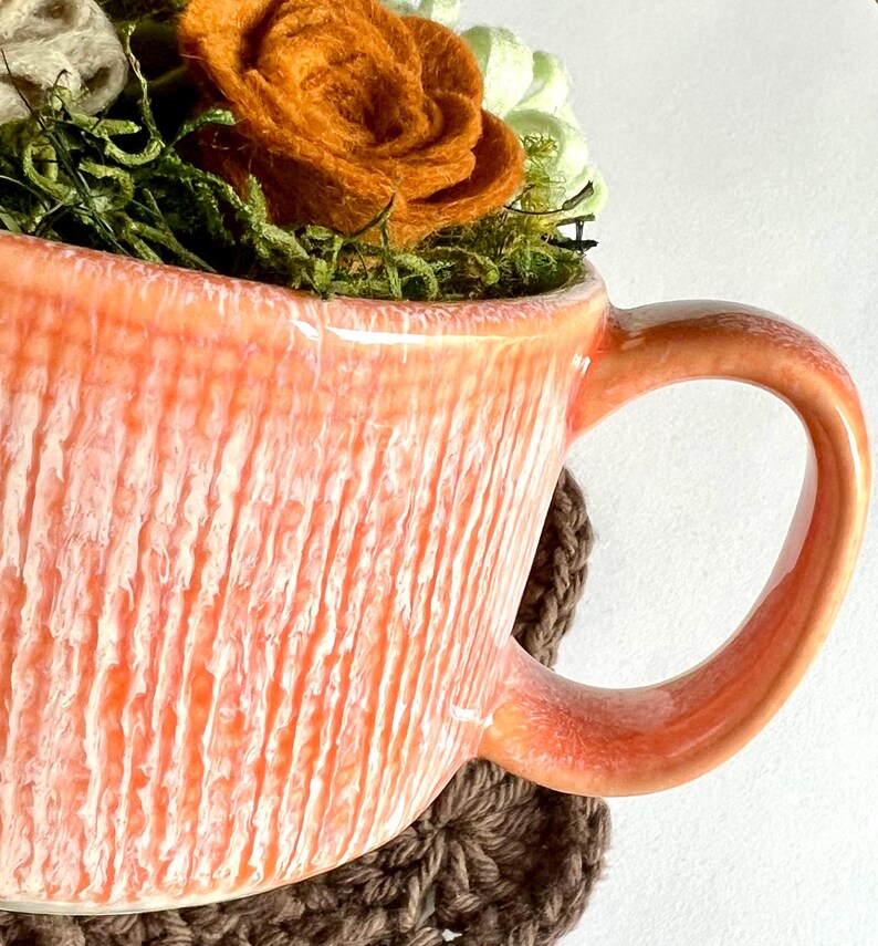 Op de afbeelding: Een perzik-kleurige keramische mok met een textuur, verticaal gestreept ontwerp. De mok is gevuld met groen mos, witte en groene vetplanten en een grote oranje vilten roos. De mok staat op een bruine gehaakte onderzetter.