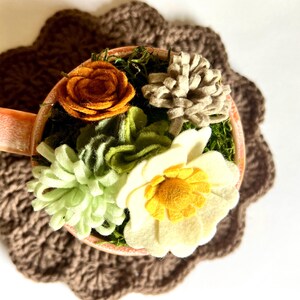 Op de afbeelding: Een decoratieve theekop gevuld met vilten bloemen in tinten geel, oranje, groen en beige, geplaatst op een bruine gehaakte onderzetter. De bloemen omvatten een roos, een madeliefje en andere bloemaccenten, waardoor een charmante, handgemaakte presentatie ontstaat.