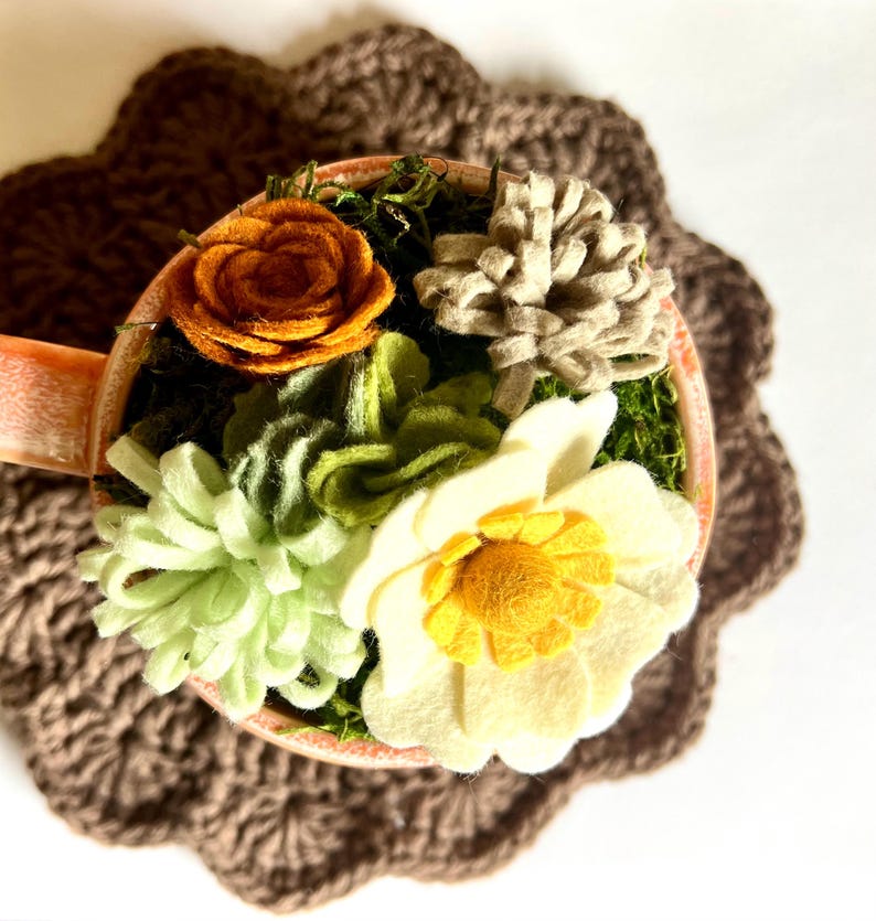 Op de afbeelding: Een decoratieve theekop gevuld met vilten bloemen in tinten geel, oranje, groen en beige, geplaatst op een bruine gehaakte onderzetter. De bloemen omvatten een roos, een madeliefje en andere bloemaccenten, waardoor een charmante, handgemaakte presentatie ontstaat.